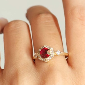 Può includere: Un anello d'oro con una pietra preziosa rossa circondata da pietre preziose più piccole e trasparenti. L'anello è su un dito.