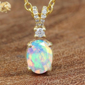 Può includere: Una collana color oro con un pendente ovale in opale. L'opale mostra una gamma di colori, tra cui blu, verde e giallo. Il pendente è impreziosito da piccole gemme trasparenti e pende da una delicata catena.