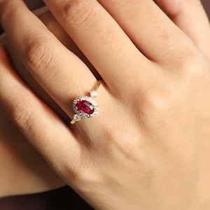 Op de afbeelding: Een gouden ring met een rode ovale edelsteen omringd door kleinere diamanten. De ring zit aan een vinger.