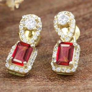 Può includere: Un paio di orecchini in oro con una pietra preziosa rossa e accenti di zircone cubico trasparente. Gli orecchini presentano un design pendente con una pietra preziosa rossa rettangolare circondata da zircone cubico trasparente.