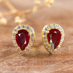 May include: A pair of gold earrings featuring pear-shaped red gemstones set within a halo of smaller white gemstones.