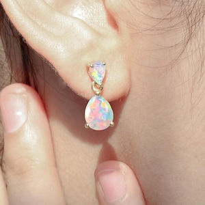 Könnte beinhalten: Ein Paar goldene Tropfenohrringe mit schillernden Opal-Steinen. Die Ohrringe hängen am Ohrläppchen.