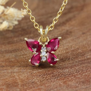 May include: A gold-coloured butterfly pendant necklace. The butterfly pendant features four pear-shaped, deep red gemstones and small white gemstones in the centre. The necklace chain is delicate and gold-coloured.