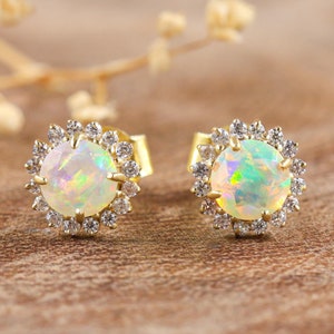 May include: A pair of gold stud earrings featuring a central opal stone surrounded by a halo of small, clear gemstones. The opals have a play of color, and the earrings are set against a warm, wooden background.