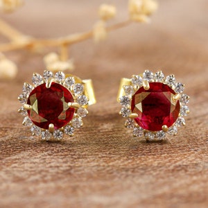 Op de afbeelding: Een paar gouden oorbellen met een rode edelsteen omringd door een halo van kleinere witte edelstenen.