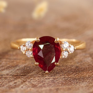 Könnte beinhalten: Ein goldener Ring mit einem birnenförmigen Rubin-Edelstein und kleineren Diamanten, die in die Band gefasst sind.