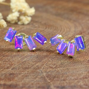 Peut inclure: Boucles d'oreilles dorées composées de cinq pierres rectangulaires, irisées, violettes et roses. Les boucles d'oreilles sont présentées sur un fond en bois, mettant en valeur les couleurs des pierres.