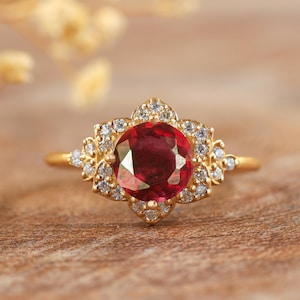 May include: A gold ring featuring a large red gemstone surrounded by smaller white gemstones.