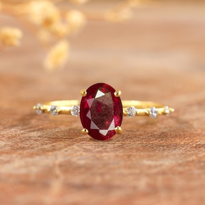 Peut inclure: Une bague en or ornée d'un grand rubis ovale. La monture est ornée de petites pierres précieuses rondes et transparentes. La bague est posée sur une surface en bois chaleureuse.