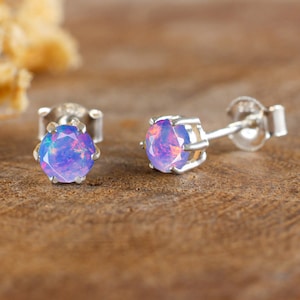 May include: A pair of stud earrings with iridescent, round, blue-purple gemstones set in silver metal. The earrings are displayed on a wooden surface, with a soft, blurred background.
