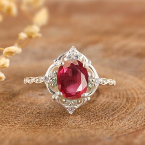May include: A silver ring featuring a large oval-cut ruby gemstone. The ruby is surrounded by a decorative setting with smaller accent stones. The ring is set against a wooden background.