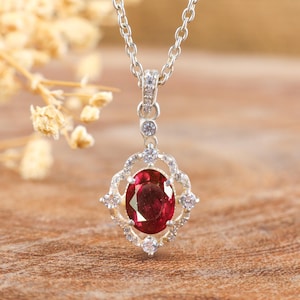 Könnte beinhalten: Eine silberne Kettenkette mit einem Anhänger, der einen großen ovalen roten Edelstein zeigt, der von kleineren klaren Edelsteinen umgeben ist.