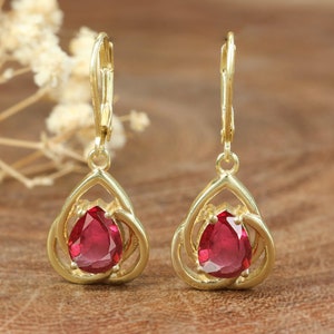 Puede incluir: Un par de pendientes de oro con una piedra preciosa roja en el centro de cada pendiente. Los pendientes tienen un diseño delicado e intrincado.