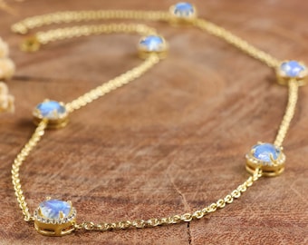 May include: A delicate gold necklace featuring multiple round, light blue gemstones. Each gemstone is set in a gold bezel and surrounded by small, clear accent stones. The necklace is displayed on a wooden surface.