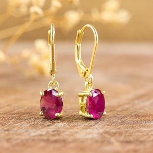 May include: A pair of gold earrings with oval-shaped ruby gemstones. The earrings have a leverback closure.