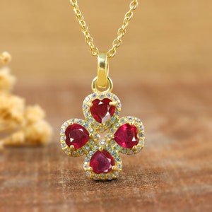 May include: A gold pendant necklace with a four-leaf clover design. Each leaf is a heart-shaped, deep red gemstone, encircled by small, clear gemstones. The necklace hangs on a delicate gold chain.