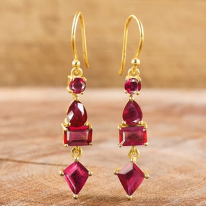 May include: A pair of gold-colored drop earrings featuring multiple red gemstones. The earrings have a hook closure and are adorned with round, pear-shaped, and square-cut stones. The gemstones are a deep red color, creating a striking contrast against the gold.