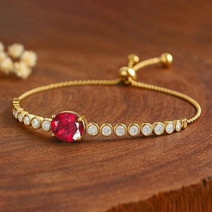 Könnte beinhalten: Ein goldfarbenes Armband mit einem großen, oval geformten, roten Edelstein in der Mitte. Das Armband ist mit kleinen, runden, klaren Edelsteinen besetzt. Der verstellbare Verschluss ist ebenfalls goldfarben.