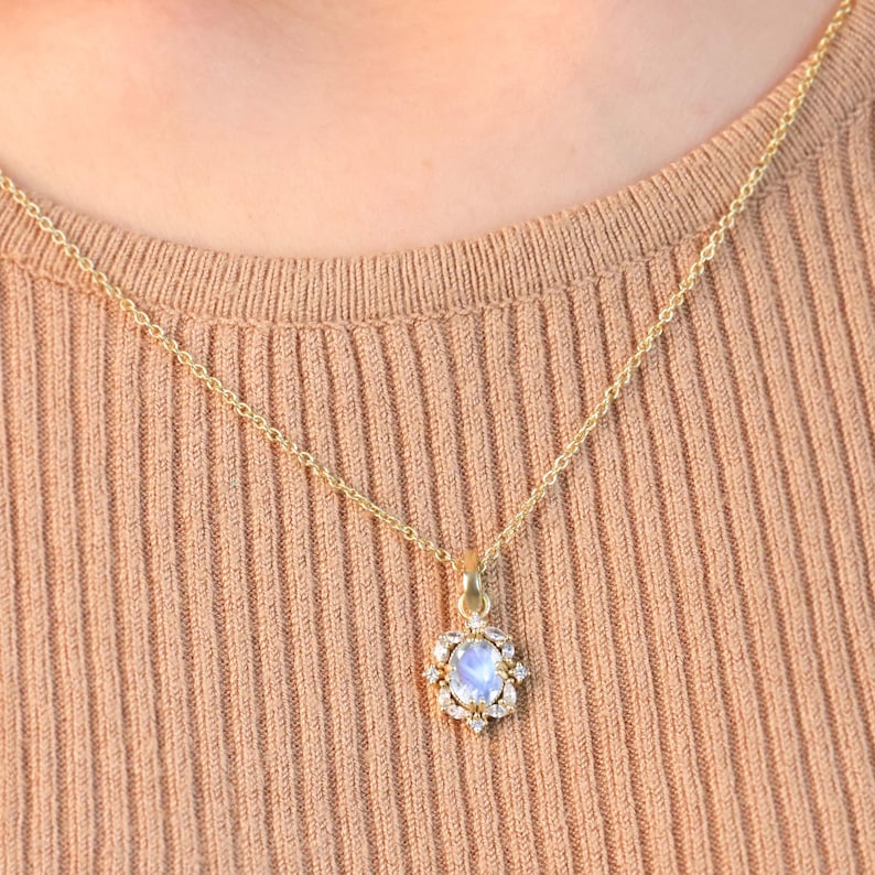 May include: A gold-colored necklace with a pendant featuring a light blue oval gemstone surrounded by small, clear stones. The pendant hangs from a delicate gold chain. The necklace is displayed against a tan ribbed fabric.