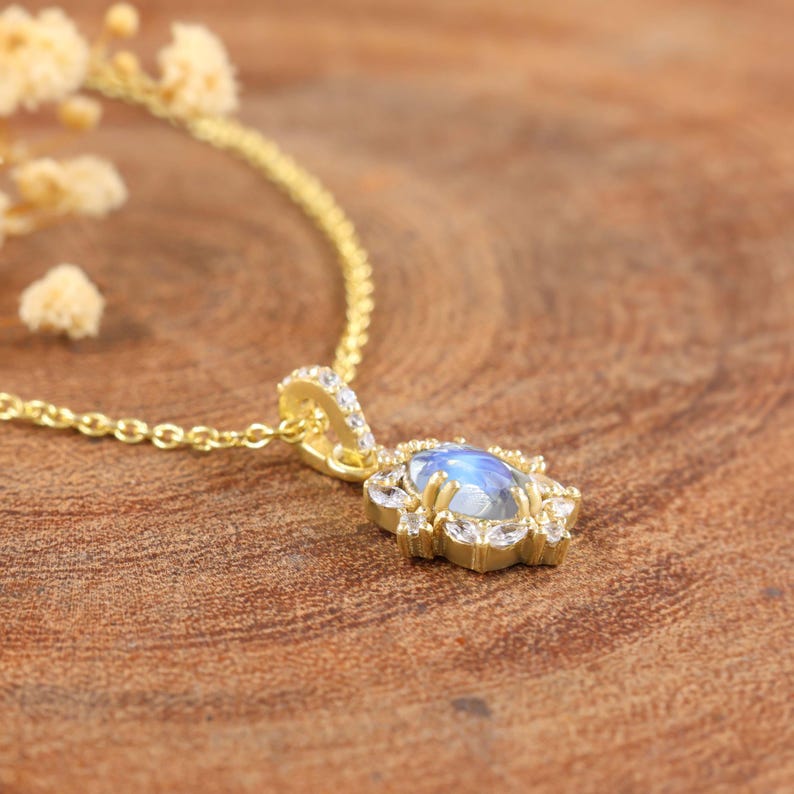 May include: A gold-colored necklace with a pendant featuring a blue gemstone surrounded by small, clear stones. The pendant is flower-shaped. The necklace is on a wooden surface.