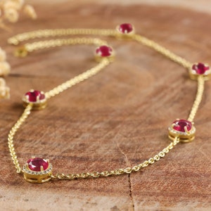 Op de afbeelding: Een gouden ketting met vijf rode robijnstenen die zijn gezet in een halo van kleinere diamanten. De ketting ligt op een bruin houten oppervlak.