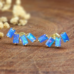 May include: A pair of gold stud earrings, each with five rectangular, iridescent blue gemstones. The gemstones display a multi-tonal effect, with flashes of pink and green. The earrings are presented on a wooden surface.