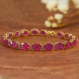 Op de afbeelding: Een gouden armband met ovale, dieprode edelstenen. De edelstenen zijn gezet in individuele gouden zettingen en met elkaar verbonden. De armband wordt gepresenteerd op een houten oppervlak, waarbij de kleur en het ontwerp worden benadrukt.