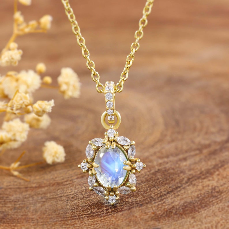 May include: A gold-colored necklace with a pendant featuring a large, oval, iridescent gemstone surrounded by smaller, clear gemstones. The pendant is suspended from a delicate chain. The necklace is set against a warm, wooden background.