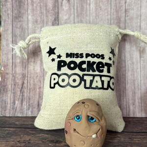 May include: A beige burlap bag with the text "MISS POOS POCKET POOTATO" and a smiling potato-shaped sculpture with blue eyes and a single tooth. The potato is brown with small brown spots.