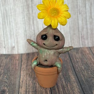 May include: A whimsical, handmade figurine of a smiling creature with a yellow flower on its head, seated in a small terracotta pot. The creature has outstretched arms and a textured, earthy appearance, with hints of green and pink. The pot sits on a wooden surface.