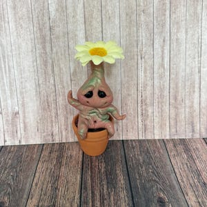 May include: A whimsical, handmade figurine of a plant-like creature with a daisy flower head. The figure is seated in a small terracotta pot. The creature has a pinkish-tan body with green accents and sad, black eyes. The background is a wooden surface.