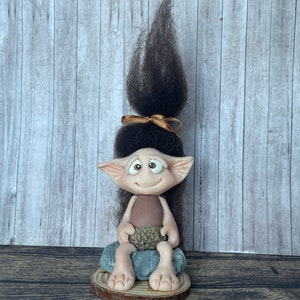 May include: A small, brown and beige troll figurine with large ears and a long, dark brown hairpiece tied with a gold ribbon. The troll is sitting on a grey rock with a brown acorn in its hand. The figurine is on a wooden base.