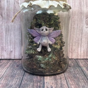 Peut inclure: Un bocal en verre transparent avec un couvercle en tissu attaché par de la ficelle, contenant une petite figurine de fée gris clair aux ailes violettes. La fée est placée devant une structure texturée vert foncé, pailletée et arborée.