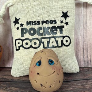 May include: A whimsical, hand-painted Pocket Pootato figurine with a smiling face and blue eyes, in front of a beige burlap bag with the text "MISS POOS POCKET POOTATO" in black. The potato has brown spots.