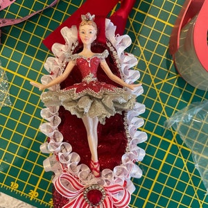 May include: A decorative ornament shaped like a ballet slipper, featuring a ballerina figurine. The slipper is red with glitter, white and red striped ribbons, and a large red jewel. The ballerina wears a red and gold tutu and a crown.