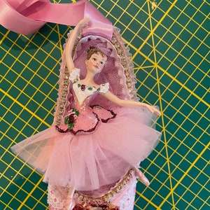 May include: A decorative pink ballet slipper ornament with a ballerina figurine. The slipper is embellished with glitter, lace, and rhinestones. A pink ribbon is attached for hanging. The ballerina is wearing a pink tutu and has her arms raised.