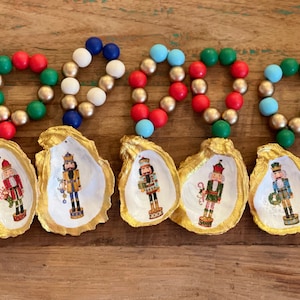 May include: Five Christmas ornaments featuring oyster shells with gold trim and nutcracker illustrations. Each shell is topped with a beaded loop in red, green, gold, white, and blue. The ornaments are displayed on a wooden surface.