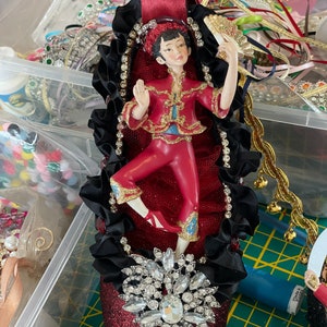 May include: A decorative shoe ornament with a red glitter finish, adorned with a figurine in a red and gold outfit. The shoe features a large, sparkling rhinestone embellishment and black ribbon accents. A burgundy ribbon extends from the top.
