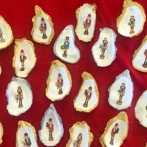 May include: A collection of oyster shells, each adorned with a gold or silver rim and a festive nutcracker design. The shells are arranged on a vibrant red surface, creating a holiday theme.