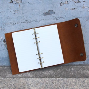Puede incluir: Un cuaderno encuadernado en cuero marrón con un inserto de papel blanco rayado y un encuadernador de anillos de latón. El cuaderno está abierto y acostado sobre una superficie gris texturizada.
