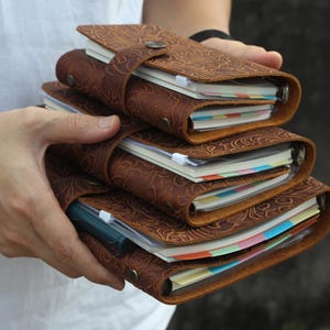 Op de afbeelding: Een stapel van vier bruine leren tijdschriften met reliëf bloempatronen. Elk tijdschrift heeft een riemsluiting en bevat kleurrijke, gelinieerde pagina's. De tijdschriften worden in handen gehouden.