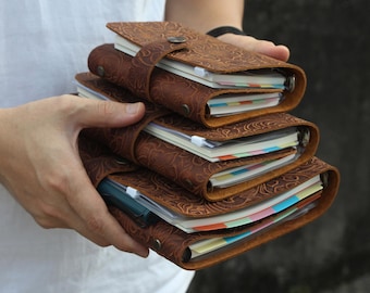 May include: A stack of four brown leather journals with embossed floral patterns. Each journal has a strap closure and contains colorful, lined pages. The journals are held in someone's hands.