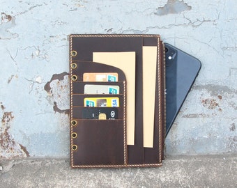 May include: A brown leather notebook with a zippered pocket and a slot for cards. The notebook has six brass rings and a brown paper insert. A black smartphone is tucked into the zippered pocket.