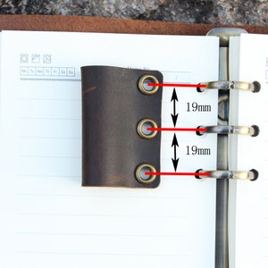 May include: A brown leather notebook holder with three brass eyelets. The eyelets are spaced 1.9 inches apart.