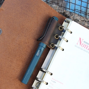 May include: A brown leather notebook cover with a pen holder and a pen. The notebook has a metal binding and the cover has six brass grommets. The notebook is open to a page with the text "Personal Information" and "個人情報" in Japanese.