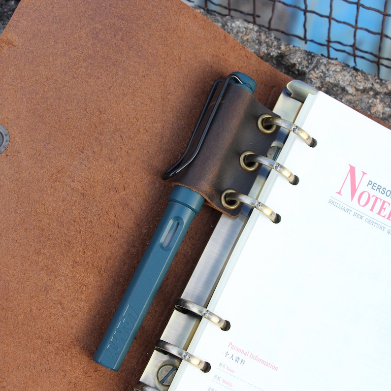 May include: A brown leather notebook cover with a pen holder and a pen. The notebook has a metal binding and the cover has six brass grommets. The notebook is open to a page with the text "Personal Information" and "個人情報" in Japanese.