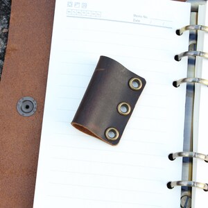May include: A brown leather notebook page holder with three brass eyelets. The holder is on a white lined notebook page.
