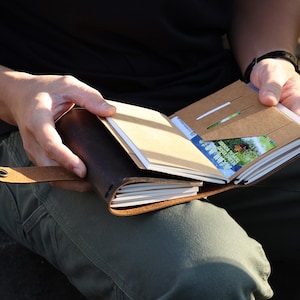 Könnte beinhalten: Ein dunkelbraunes Lederjournal mit Druckknopfverschluss. Das geöffnete Notizbuch enthält mehrere weiße Papiereinlagen, ein beiges Innenfach und eine kleine rechteckige Karte mit blau-grünen Motiven, die in das Fach gesteckt ist.
