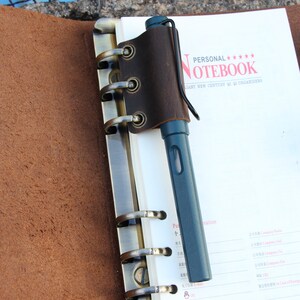 May include: A brown leather notebook with a pen holder and a metal ring binder. The notebook has the text "PERSONAL***** NOTEBOOK" and "BRILLIANT NEW CENTURY QI QI ORGANIZERS" printed on the cover.