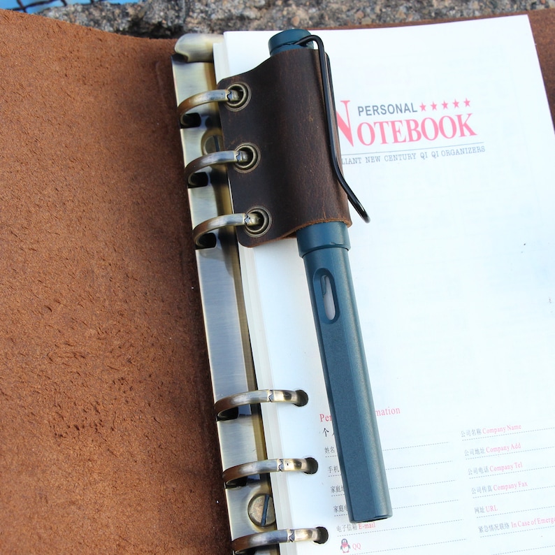 May include: A brown leather notebook with a pen holder and a metal ring binder. The notebook has the text "PERSONAL***** NOTEBOOK" and "BRILLIANT NEW CENTURY QI QI ORGANIZERS" printed on the cover.